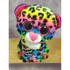 May include: A plush leopard toy featuring a vibrant rainbow-colored fur pattern with black spots. The stuffed animal has large, round, glittery dark eyes, a bright pink nose, and a white muzzle area. The ears are purple with pink inner lining.