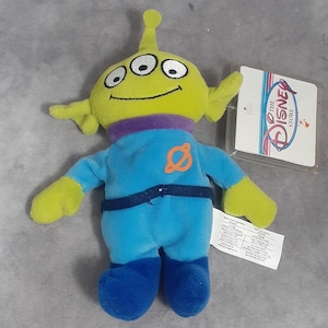 May include: A plush toy alien from the Disney Store, featuring a lime green head with three eyes, yellow arms, and a blue jumpsuit. The alien has a purple neck and blue boots. A Disney Store tag is attached.