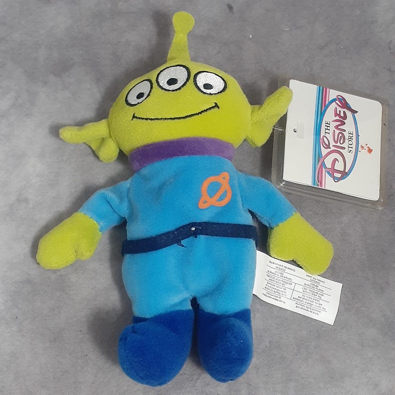 May include: A plush toy alien from the Disney Store, featuring a lime green head with three eyes, yellow arms, and a blue jumpsuit. The alien has a purple neck and blue boots. A Disney Store tag is attached.