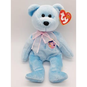 May include: A light blue Beanie Baby bear with a pink ribbon around its neck. The bear has a small embroidered patch on its chest that says "ty" and has a colorful design. The bear is sitting on a white background.
