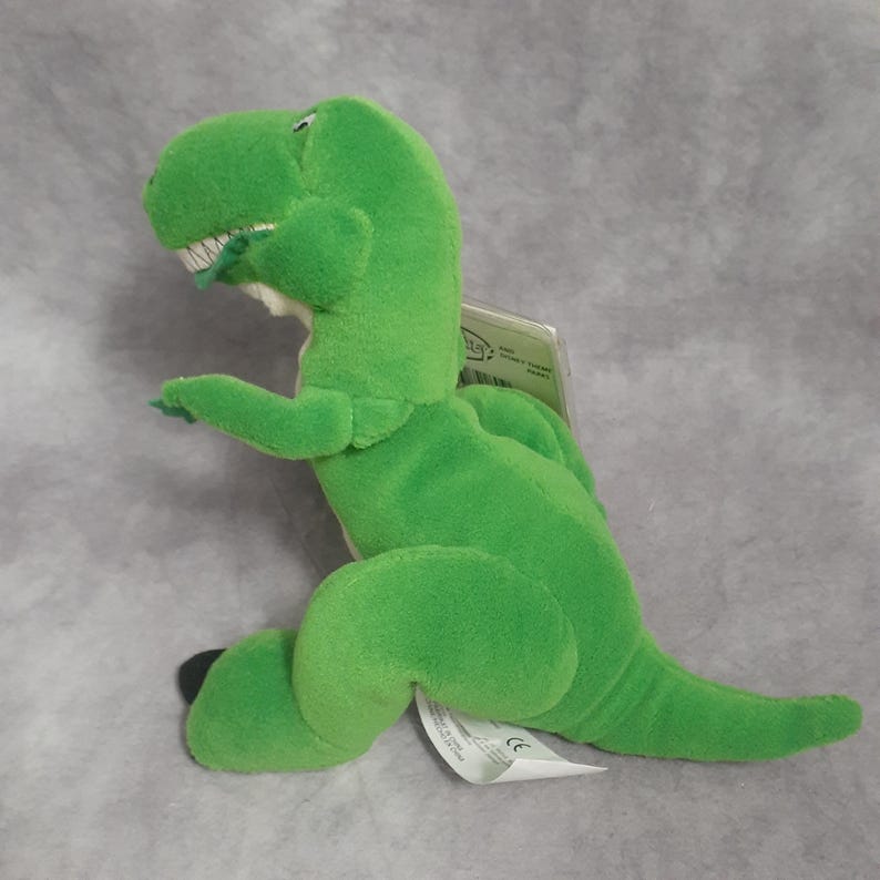 May include: A plush green Tyrannosaurus Rex toy with white teeth and a tag attached. The dinosaur is in a sitting position, with its arms and legs extended. The toy is made of soft material.