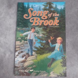 May include: A book cover titled "Song of the Brook, Second Edition" featuring a scenic illustration. The image depicts a boy and a girl near a stream, with a butterfly. The book is part of "A Beka Book Reading Program."