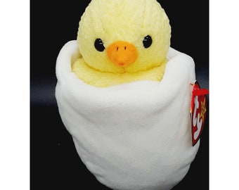 May include: A yellow chick plush toy in a white egg-shaped plush. The chick has black eyes, an orange beak, and a white beanie. The toy has a red and white Ty tag.