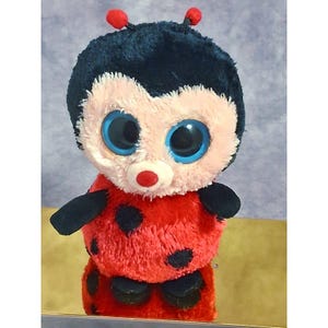 May include: A plush ladybug toy featuring a fuzzy red body with black spots, a light pink face, and large, round, bright blue eyes. The toy has a black head with two small antennae topped with red spheres and small black arms and feet.