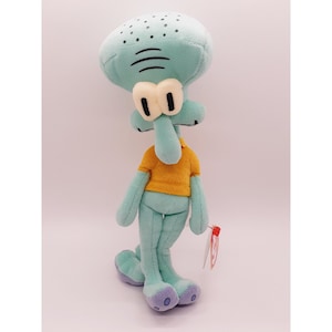 May include: A green and purple plush toy of Squidward Tentacles from the cartoon Spongebob Squarepants. He is wearing a yellow shirt and has a white tag attached to his leg.