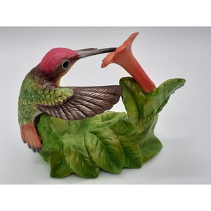 May include: A porcelain figurine of a hummingbird with green and red feathers, perched on a green leafy branch with a red flower. The hummingbird is depicted with its wings spread, as if in flight.