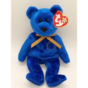 May include: Blue Beanie Baby teddy bear with a gold ribbon and a gold embroidered European Union symbol. The bear has a red Ty Beanie Babies tag attached to its ear.