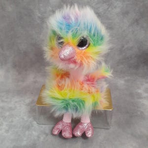 May include: A colorful plush bird with rainbow-colored fur, a pink glittery beak, and matching glittery feet. The bird has large black eyes and is sitting on a small, silver box. The toy is a whimsical and vibrant creation.