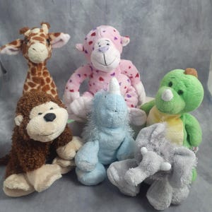 May include: A collection of six plush animal toys. The toys include a giraffe with brown spots, a pink monkey with heart patterns, a brown monkey, a blue unicorn, a green dinosaur, and a gray elephant.