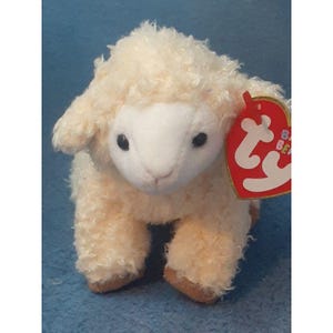 May include: A white plush lamb with a brown nose and black eyes. The lamb is sitting on a blue surface and has a red Ty Beanie Baby tag attached to its ear.