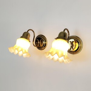 May include: Two brass wall sconces with white ruffled glass shades. The sconces have a traditional design and are illuminated with warm light.