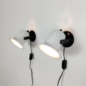 May include: Two white and black wall-mounted spotlights. Each spotlight has a white, cone-shaped shade and a black mounting plate. The lights are angled downward, with a black cord and switch. The lights are on.