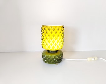 Könnte beinhalten: Eine Vintage-Tischlampe mit gelbem Glasschirm und grünem Sockel. Der Schirm hat ein Rautenmuster und der Sockel ein strukturiertes Design. Die Lampe ist mit einem weißen Kabel und einem Schalter angeschlossen.