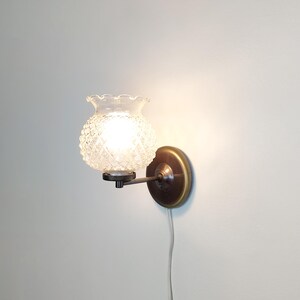 Può includere: Un'applique vintage con un paralume in vetro trasparente e strutturato e una base in metallo color ottone. La luce è accesa, illuminando il vetro a motivo diamantato. L'applique è montata su una parete bianca.