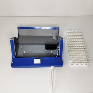 May include: A blue metal box with a hinged lid, open to reveal a grey interior and a black cylindrical component. A white grid-like structure sits to the right. White cords extend from the box.