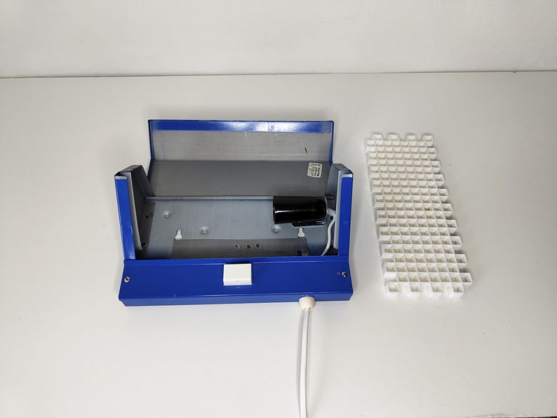 May include: A blue metal box with a hinged lid, open to reveal a grey interior and a black cylindrical component. A white grid-like structure sits to the right. White cords extend from the box.