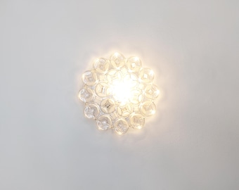 May include: A round, clear glass light fixture with a bright central light source. The fixture is composed of multiple clear, circular glass elements arranged in a floral pattern. The light fixture is mounted on a white wall.