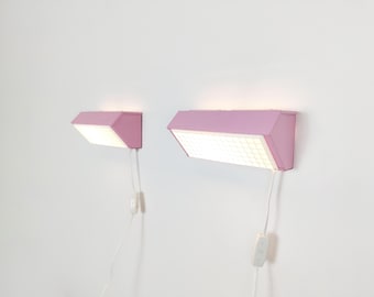 Puede incluir: Dos lámparas de pared con carcasas de metal rosa claro y difusores de luz rectangulares con patrón de rejilla. Cada lámpara cuenta con un cable de alimentación blanco con interruptor integrado, emitiendo una luz suave y cálida.