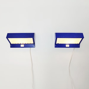 May include: Two blue wall-mounted lamps with a rectangular design. Each lamp has a grid of small lights behind a blue cover. White cords extend from the bottom of each lamp. The lamps are mounted on a white wall.