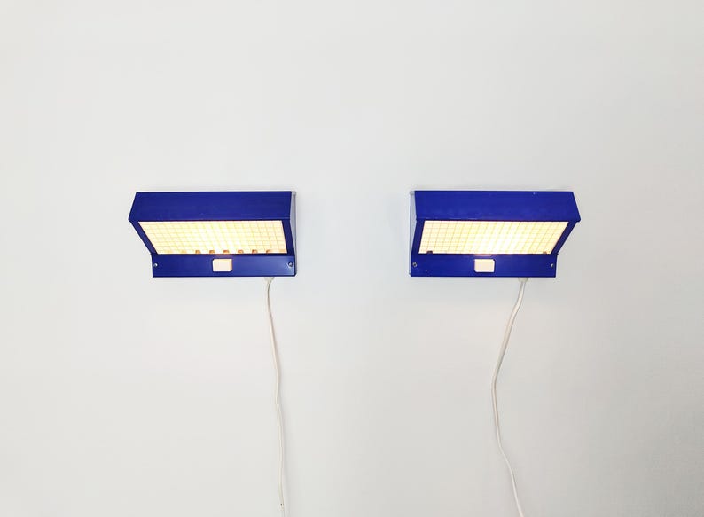 May include: Two blue wall-mounted lamps with a rectangular design. Each lamp has a grid of small lights behind a blue cover. White cords extend from the bottom of each lamp. The lamps are mounted on a white wall.