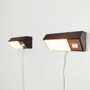 May include: Two wall-mounted lamps featuring dark brown metal housings, rectangular light panels with a grid pattern, and wood-grain accents. Each lamp includes a white power switch and a white electrical cord extending downward from the base.