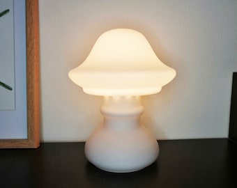 Könnte beinhalten: Eine weiße Keramik-Tischlampe in Pilzform mit einem runden, mattierten Glasschirm. Die Lampe ist eingeschaltet und strahlt ein warmes, weißes Licht aus.