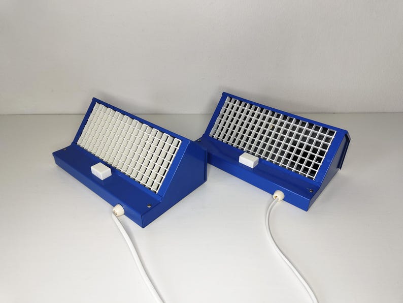 May include: Two blue desk lamps with white gridded diffusers. Each lamp has a white switch and a white cord. The lamps are angled and sit on a white surface.