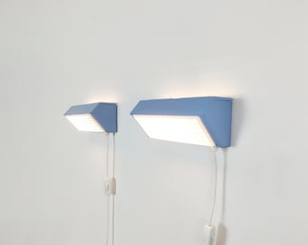 May include: Two light blue wall-mounted lamps with angled, rectangular housings and white, grid-patterned diffusers. Each lamp features a white power cord extending downward, equipped with an inline rocker switch for manual operation.