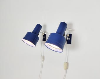 Puede incluir: Dos lámparas de pared de metal azul con cabezales cilíndricos e interiores blancos. Cada lámpara cuenta con un pequeño soporte rectangular, un cable de alimentación blanco con interruptor y una fuente de luz brillante en la pantalla.