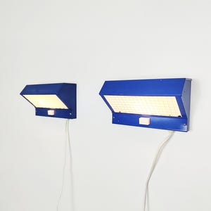May include: Two blue wall lamps with a rectangular, grid-like light panel. Each lamp has a switch and a white cable. The lamps are mounted on a white wall, offering a modern lighting solution.