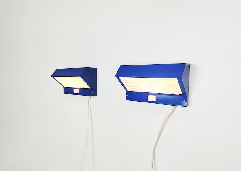 May include: Two blue wall lamps with a rectangular, grid-like light panel. Each lamp has a switch and a white cable. The lamps are mounted on a white wall, offering a modern lighting solution.