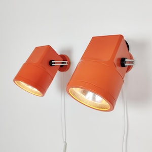 May include: Two orange wall-mounted spotlights with a cylindrical head and a square base. Each lamp features a silver-toned metal hinge mechanism on the side, a white pull cord, and a warm light emitting from the circular opening at the front.
