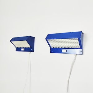 May include: Two blue wall-mounted lamps with a grid of white squares. The lamps have a rectangular shape with a slanted top and a white cord. The lamps are mounted on a white wall.