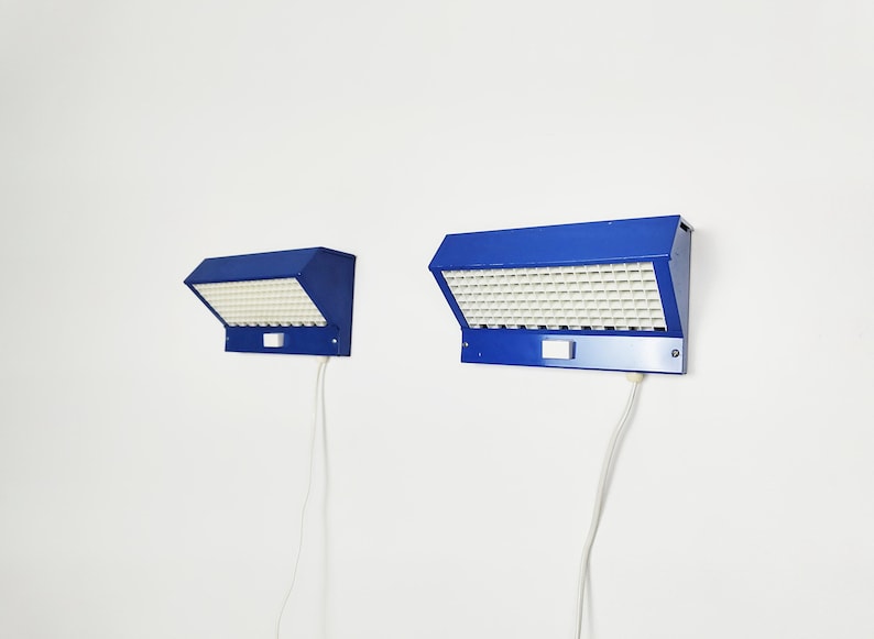 May include: Two blue wall-mounted lamps with a grid of white squares. The lamps have a rectangular shape with a slanted top and a white cord. The lamps are mounted on a white wall.