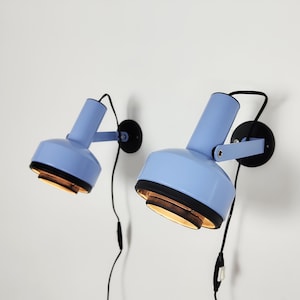 Può includere: Una coppia di lampade da parete color azzurro con teste cilindriche, piastre circolari nere e bracci regolabili. Ogni lampada presenta un bordo nero attorno all'apertura della luce, un cavo nero e un piccolo interruttore sul filo.