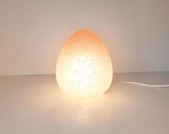 Könnte beinhalten: Eine eiförmige Lampe mit strukturierter Oberfläche, die ein warmes, weiches Leuchten ausstrahlt. Das Licht geht von einer hellen Mitte zu einem helleren Orange an der Oberseite über. Ein weißes Netzkabel erstreckt sich von der Basis.