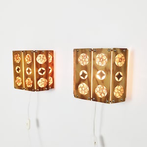 May include: Two rectangular wall sconces made of polished brass with a warm metallic finish. Each fixture features three vertical panels with circular cutouts containing intricate wire coil patterns and diamond-shaped openings, emitting a soft light.