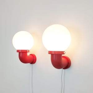 Può includere: Due lampade da parete con globi. Ogni lampada ha un paralume a globo bianco brillante sopra un raccordo per tubi rosso curvo. Gli apparecchi sono montati su una parete bianca, con un cavo bianco che pende da ogni lampada.