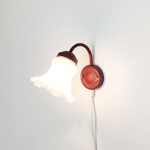 Könnte beinhalten: Eine Wandleuchte mit einem dunkelroten Metallsockel und einem geschwungenen Arm, der einen leuchtenden, gewellten weißen Glasschirm trägt. Die Leuchte verfügt über ein weißes Stromkabel, das von der runden Montageplatte nach unten führt.