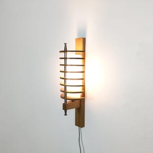 Könnte beinhalten: Eine Wandleuchte mit einer vertikalen Holzrückplatte und einem zylindrischen weißen Glasschirm. Der Schirm ist von einer Reihe horizontaler Holzlamellen umgeben, die von einer zentralen Metallstange mit Endstücken zusammengehalten werden.