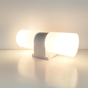May include: A white, modern wall-mounted light fixture with two cylindrical, frosted glass shades. The fixture has a curved arm and a rectangular base, casting a warm glow.