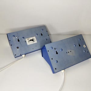 May include: Two blue metal wall sconces with white cords. The sconces have a rectangular shape with a blue front and a light blue back. Each sconce has a sticker with a black and white image of a horse.