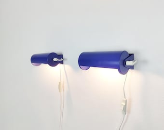 Puede incluir: Dos lámparas de pared cilíndricas con un acabado en azul vibrante, cada una con un cable blanco y un interruptor integrado. Las lámparas emiten una luz cálida desde sus extremos abiertos, mostrando un diseño tubular montado en la pared.