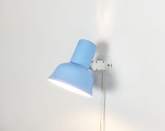 Puede incluir: Una lámpara de pared de color azul claro con una pantalla en forma de campana y una base blanca. La lámpara está fijada a una pared blanca e incluye un cable de alimentación blanco que baja desde el soporte, iluminando desde la pantalla.