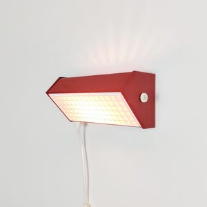 Könnte beinhalten: Eine rote Wandleuchte mit einem rechteckigen, abgewinkelten Gehäuse und einem weißen Diffusor mit Gittermuster. Die Leuchte strahlt ein warmes, helles Licht von der Vorderseite ab und hat einen kleinen weißen runden Schalter an der Seite sowie ein Kabel.