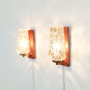 Può includere: Due applique da parete con piastre posteriori rettangolari in legno e bracci in metallo color ottone. Ogni lampada presenta un paralume in vetro trasparente testurizzato con un motivo ovale ripetuto che diffonde la luce interna.