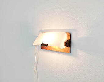 Puede incluir: Un aplique de pared con una placa trasera rectangular de madera y una pantalla curva de vidrio blanco esmerilado. La lámpara incluye una pequeña pieza de madera como detalle en el centro y un cable de alimentación blanco en el lado izquierdo.