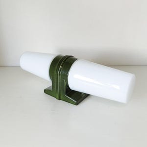 Könnte beinhalten: Eine Wandleuchte mit einem dunkelgrünen Keramiksockel in gestuftem, geometrischem Design. Zwei zylindrische, undurchsichtige weiße Glasschirme ragen horizontal aus der Mitte heraus und erzeugen ein ausgewogenes, symmetrisches Erscheinungsbild.