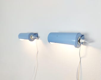 Puede incluir: Dos lámparas de pared cilíndricas de color azul claro montadas horizontalmente. Cada lámpara presenta un cuerpo de tubo metálico, un cable eléctrico blanco que sale por el lateral y una fuente de luz que emite un brillo cálido desde la base.