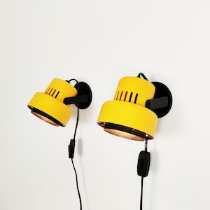 May include: Two yellow wall-mounted lamps with black accents and a simple design. The lamps have a round shape and a small switch on the cord.